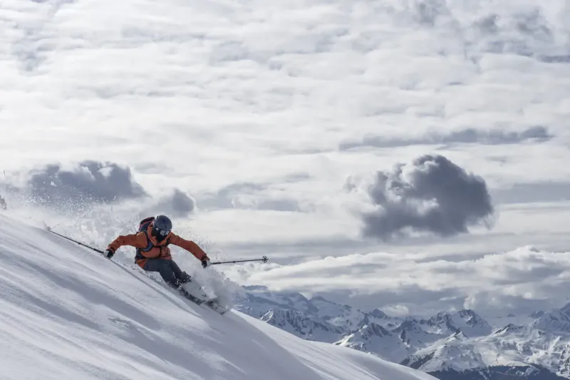 Skier in orange jacket and blue helmet carves down snowy slope in Val d'Aran with mountains and clouds