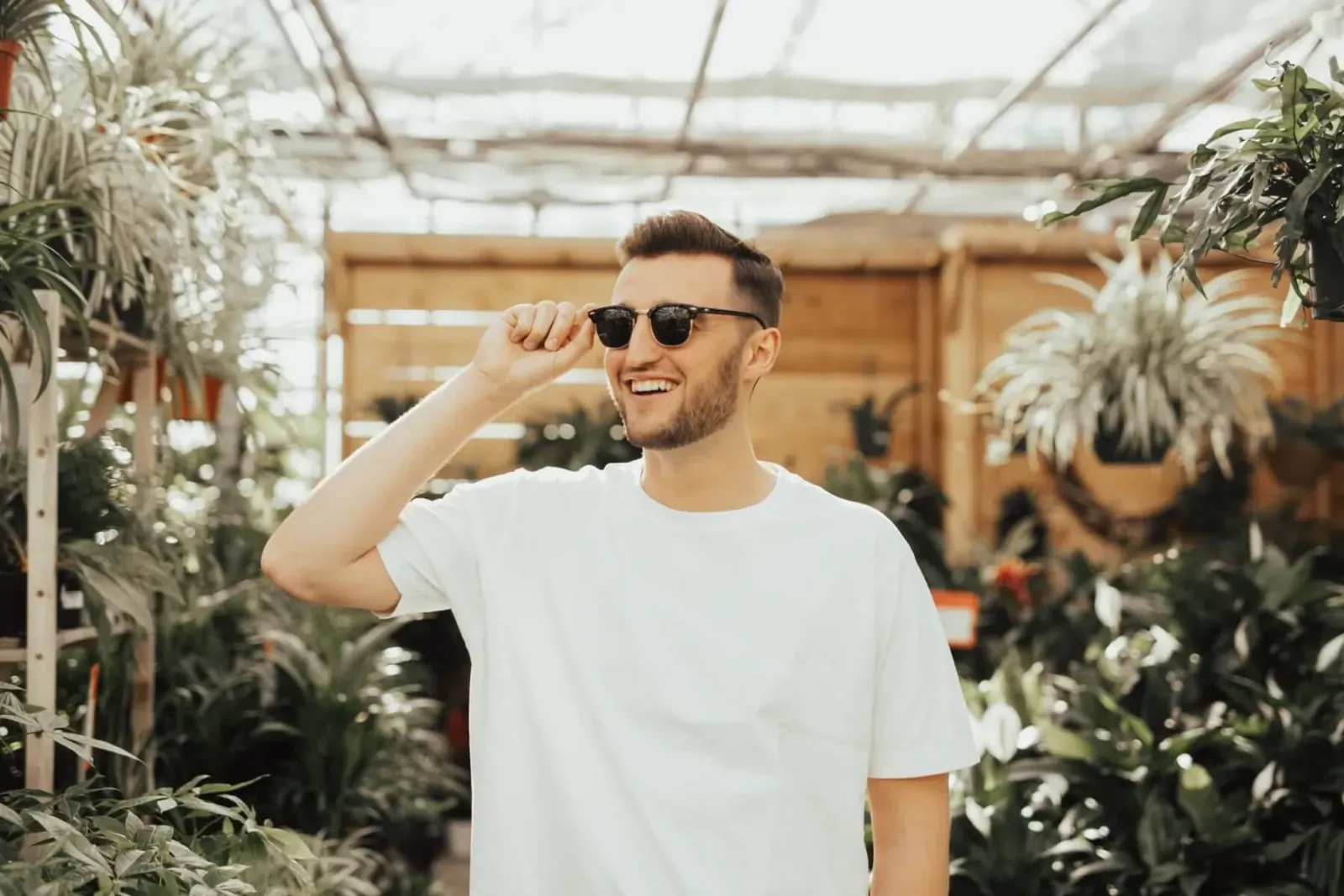 Smiling man with short hair and sunglasses adjusts shades in lush greenhouse, wearing white graphic tee