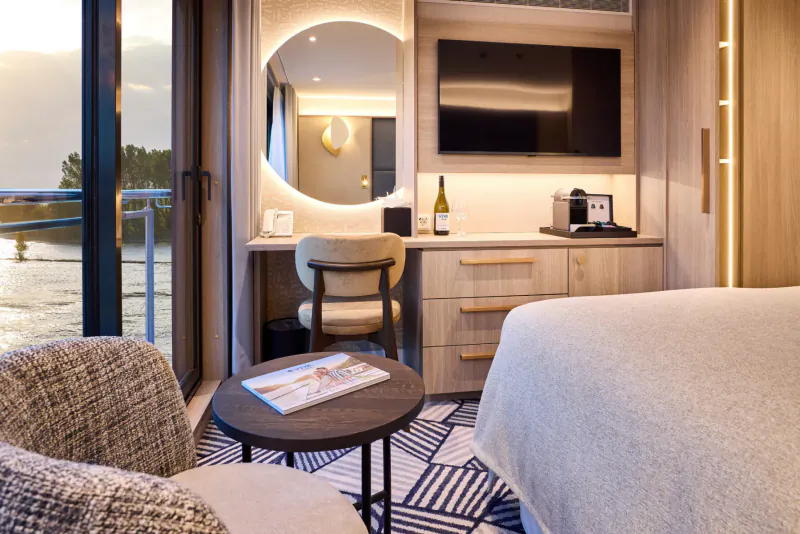 Luxurious river cruise cabin with desk, chair, bed, TV, Nespresso, wine bottle, balcony overlooking river at sunset.