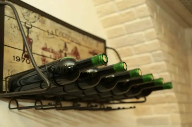 Wall-mounted wine rack with 6 green-corked bottles on beige brick wall, sign reads 'LA CORBEILLE 1970' with deer motif.