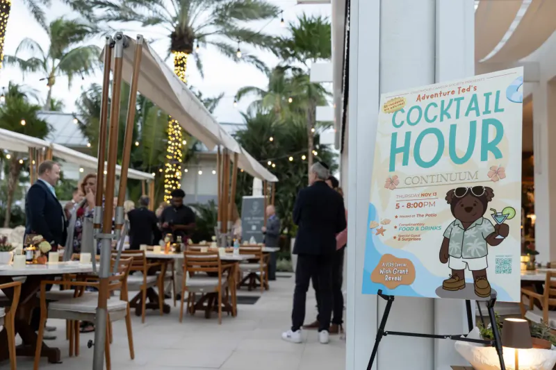 Outdoor patio dining with palm trees and string lights; 'Adventurer's Club Cocktail Hour' poster featuring cartoon bear in Hawaiian shirt.