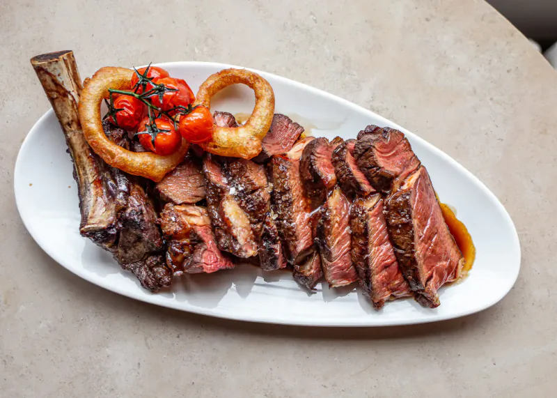 Oval white plate with sliced ribeye steak on bone, topped with onion rings and cherry tomatoes