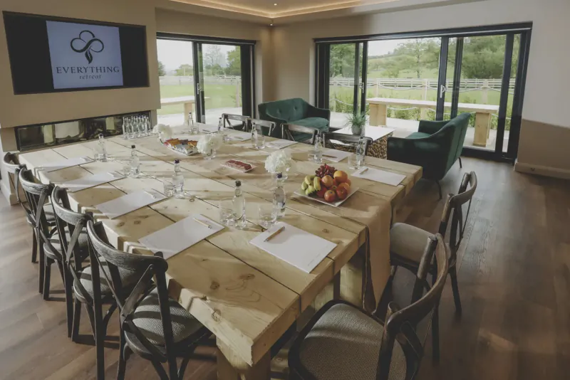 Spacious wooden conference table set with water, fruit bowls, and papers in Everything Retreat room with garden view.