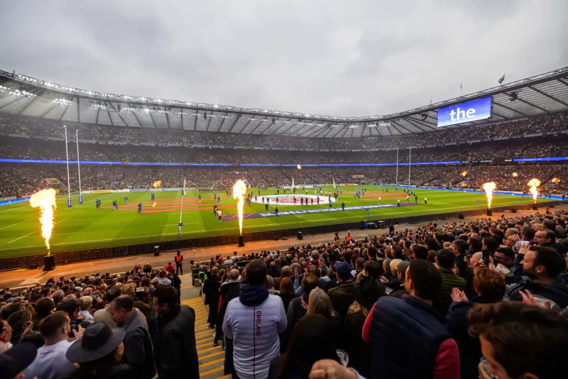 Packed rugby stadium with flaming flares on pitch during match, cheering crowds under stadium lights