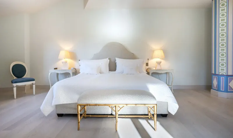 Elegant hotel bedroom with white upholstered bed, gold bench, blue chair, lamps, and ornate wall tiles.
