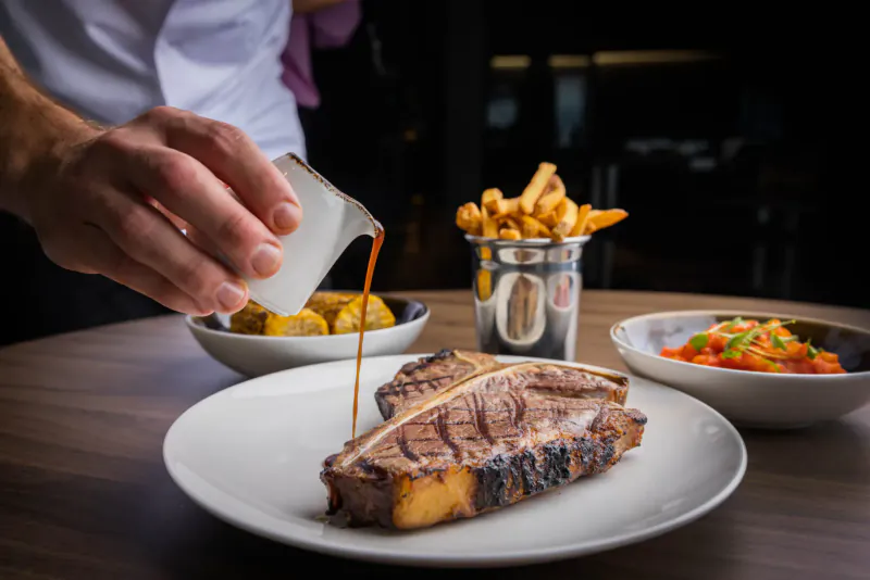 Chef pouring sauce over grilled T-bone steak on plate with fries, yuca fries, and tomato salad