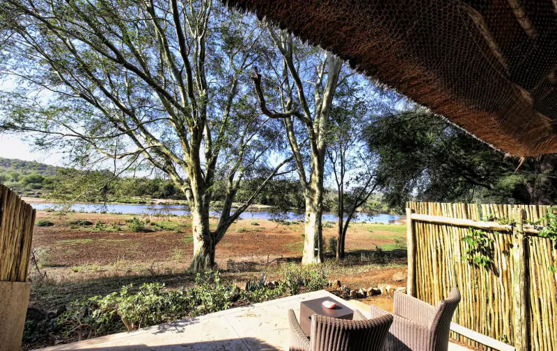 Outdoor deck of bush lodge with chairs overlooking river, acacia trees, and savanna landscape.