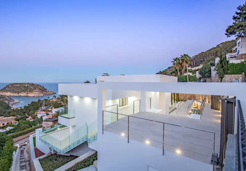 Modern white luxury villa terrace at dusk, overlooking sea bay and hillside homes in Spain.