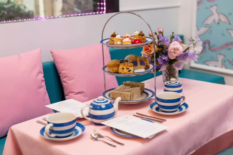 Three-tier stand with cakes, scones, and pastries on pink table with blue-striped teacups, teapot in vibrant cafe.