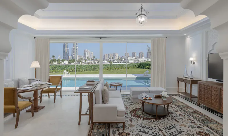 Luxurious hotel suite living room with white sofas, ornate rug, pool, and Dubai skyline view through large windows.