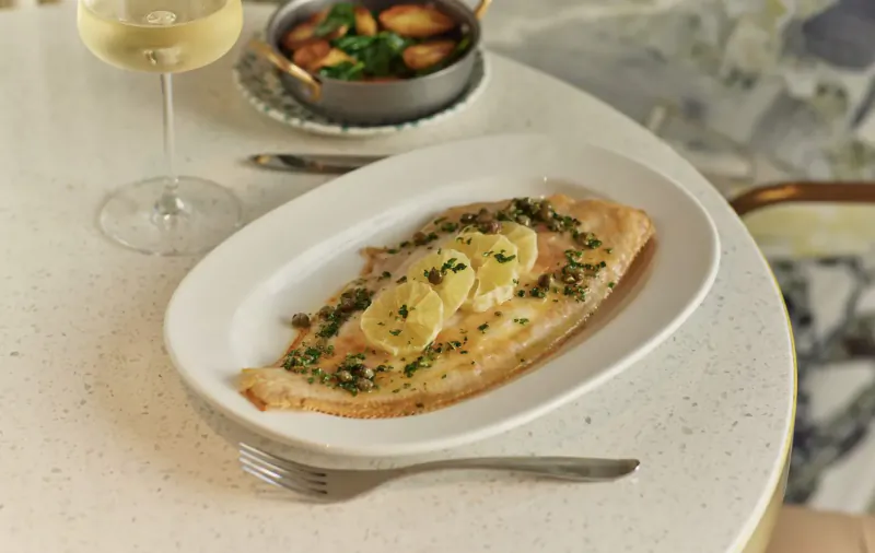 Oval white plate with lemon-caper sole fillet, garnished with lemon slices, beside white wine glass and side of roasted potatoes on marble table at Park Corner Brasserie.
