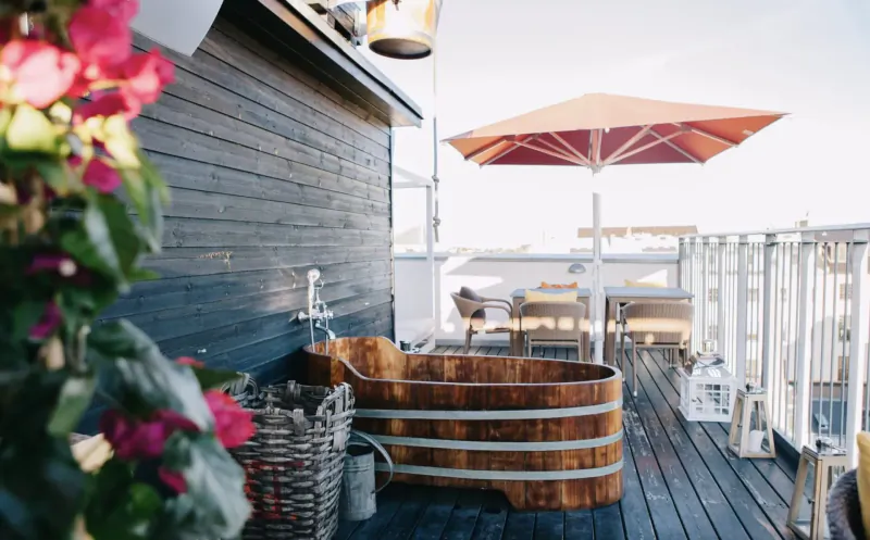 Wooden hot tub on sunny rooftop deck with red umbrella, pink flowers, table, chairs, and ocean view at Manon Les Suites.