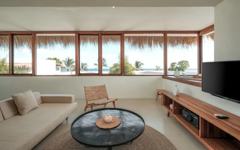 Spacious beach resort living room with rattan chair, beige sofa, round black table, wood media unit, and ocean views through thatched windows.