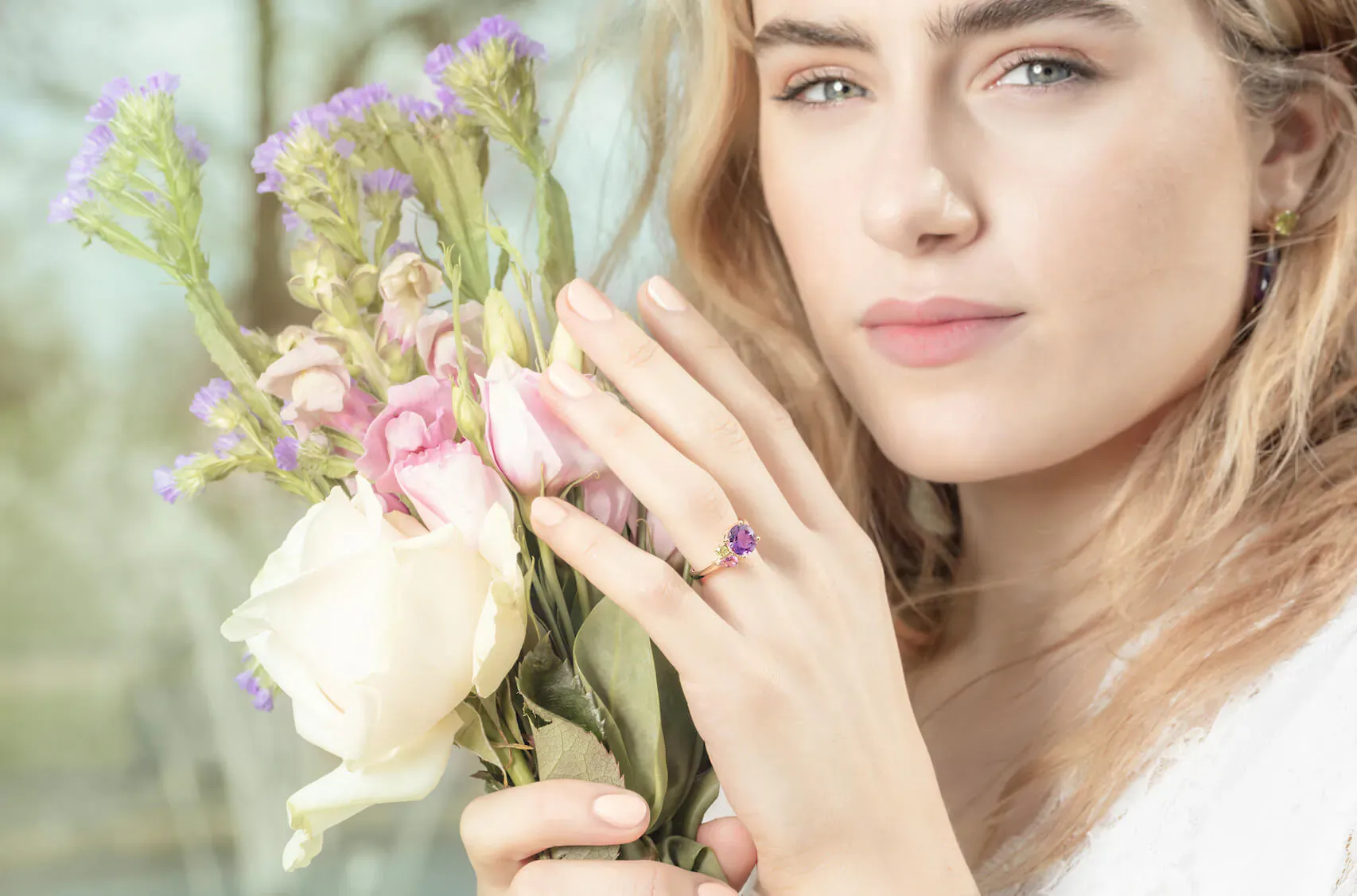 Blonde woman in white holds bouquet of pink roses, lavender flowers, and white rose, showcasing purple gem ring on finger