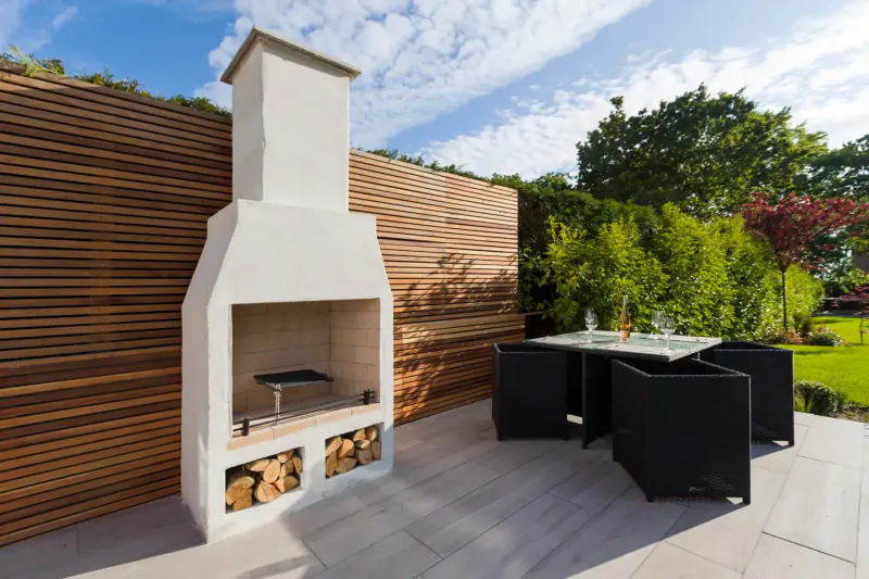 Schiedel Isokern Volcanic white garden fireplace with firewood, beside black outdoor dining table on patio amid greenery