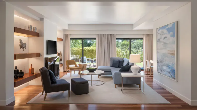 Spacious modern living room in Sheraton Cascais Resort with gray sofas, wood floors, large windows to garden, and abstract art.
