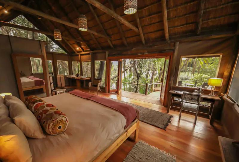 Luxurious thatched lodge bedroom with king bed, desk, and sliding doors to Okavango Delta deck view.