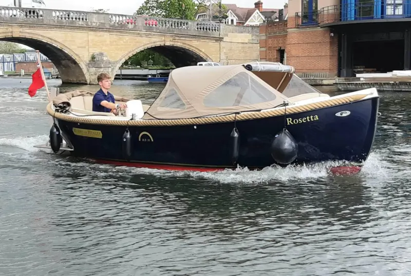 Man steers blue motorboat 'Roseveare' with beige canopy under arched bridge on river, UK flag flying.