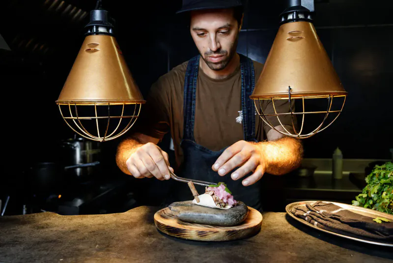 Chef David Rivas in denim apron slicing garnished dish on stone plate under hanging lamps in kitchen