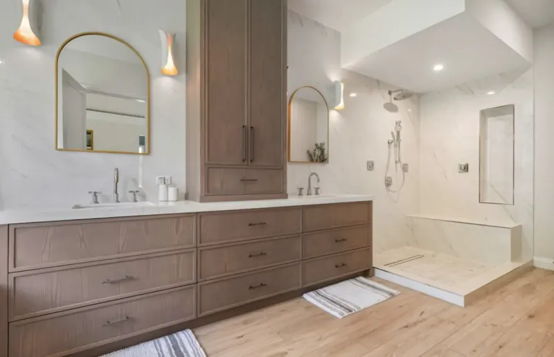 Modern luxury bathroom with double wood vanity, arched mirrors, white marble, glass shower, and wood floors.