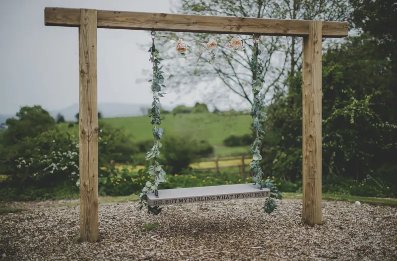 Rustic wooden swing bench draped in ivy and flowers, hanging in garden with green hills backdrop
