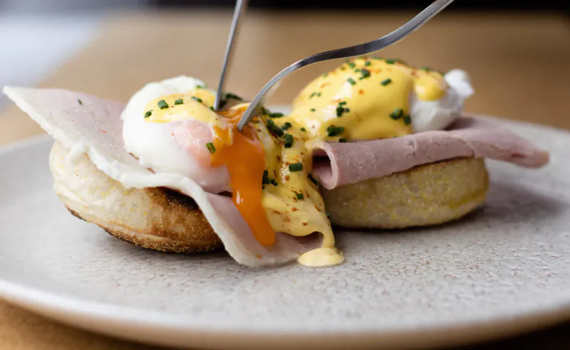 Close-up of eggs Benedict on English muffins with ham, poached eggs, hollandaise sauce, and chives, fork lifting a bite.