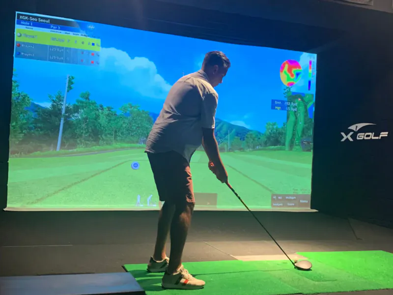 Man in white shirt and shorts swings golf club on X Golf simulator with virtual course on large screen