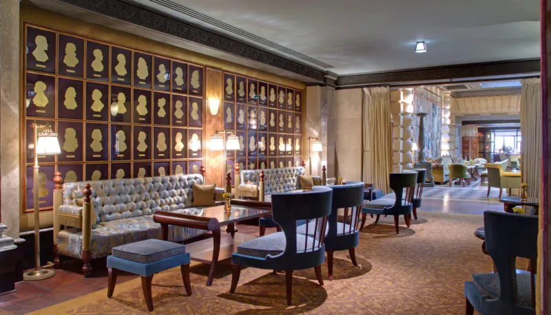 Elegant Hotel Metropole Monte-Carlo lounge with gold-framed silhouette portraits, tufted sofas, armchairs, lamps, and warm carpeted floor.