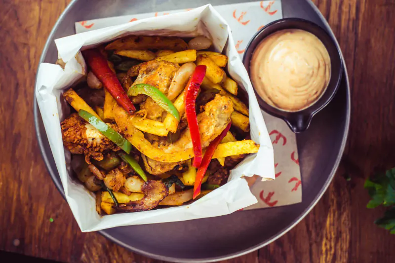 Chicken and fries with bell peppers in paper box, served with dipping sauce on wooden table