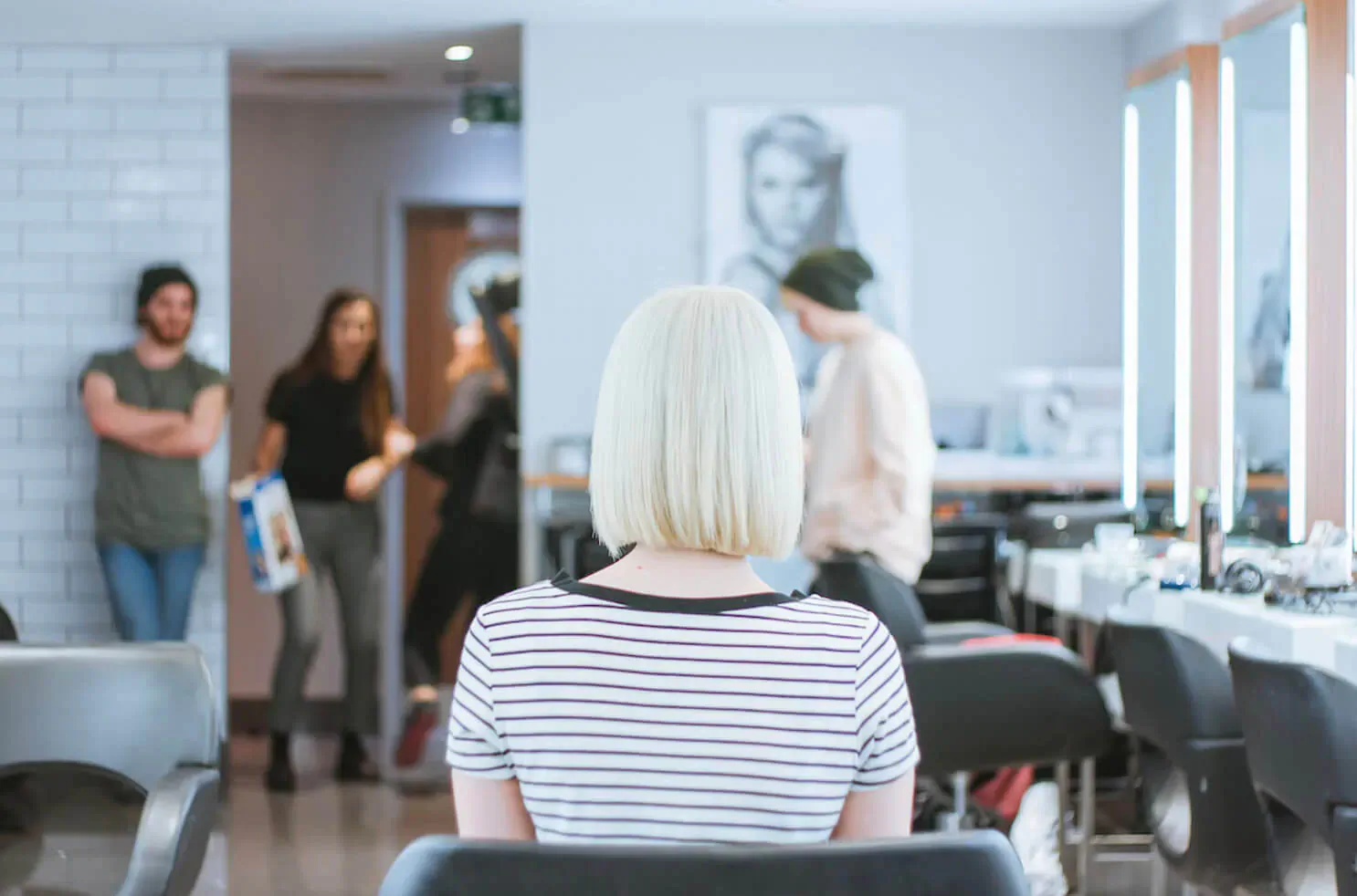 Blonde woman with bob haircut seated at salon chair, viewed from behind, with stylists and mirrors in modern hair salon