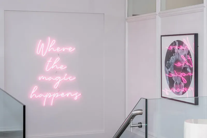 Pink neon sign 'Where the magic happens' beside framed artwork near glass staircase in stylish clinic gallery