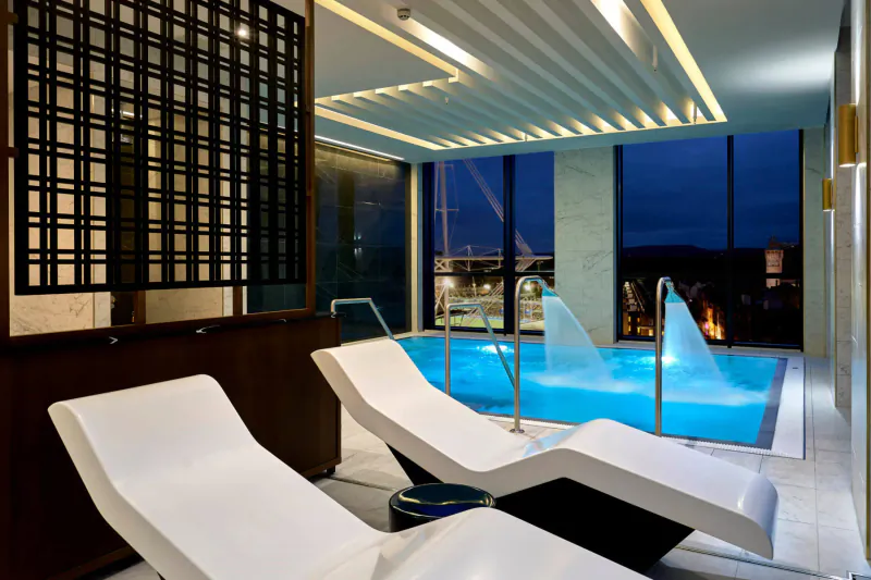 Luxurious indoor pool with glowing blue water, cascading jets, white loungers, and city night view through windows.