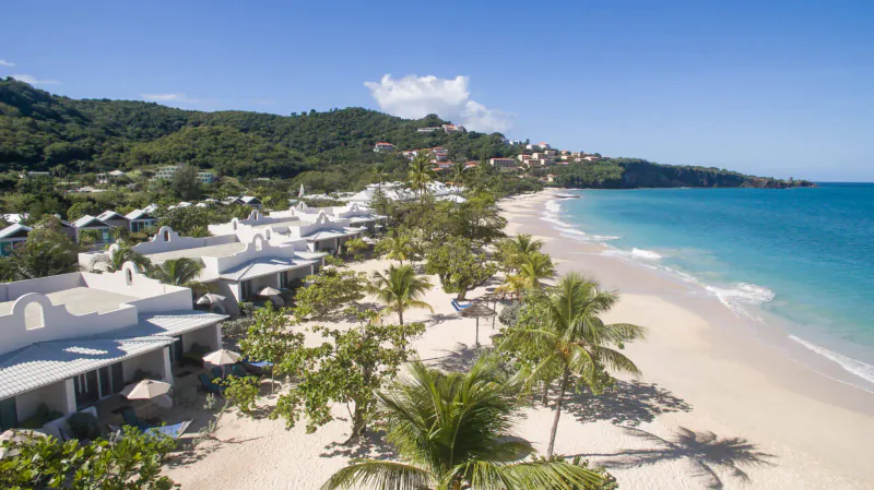 Aerial view of Spice Island Beach Resort in Grenada: white villas line a palm-fringed white-sand beach with turquoise sea and hills.