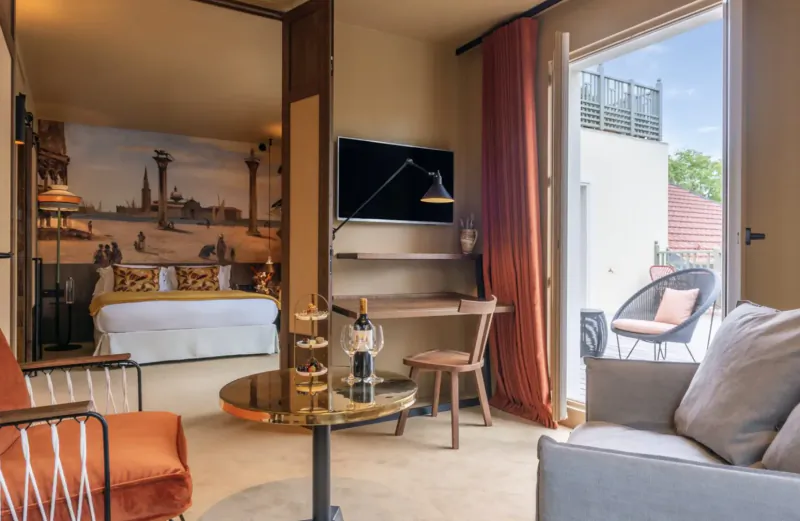 Luxurious hotel suite with Eiffel Tower mural, king bed, orange armchairs, desk, wine bottle on table, open balcony to garden.
