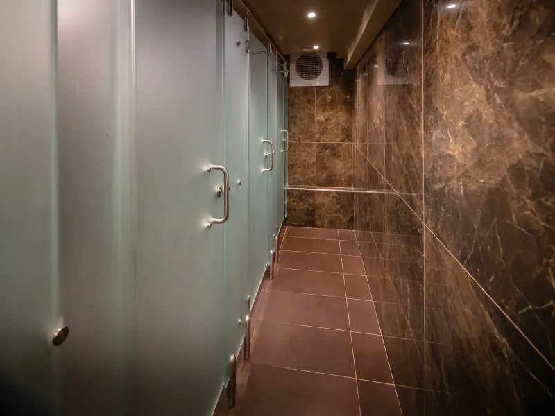 Modern luxury hotel restroom with frosted glass stalls, marble walls, brown tile floor, and ceiling lights.
