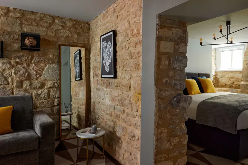Cozy stone-walled hotel room with yellow sofa, round table, mirror, art, and bed.