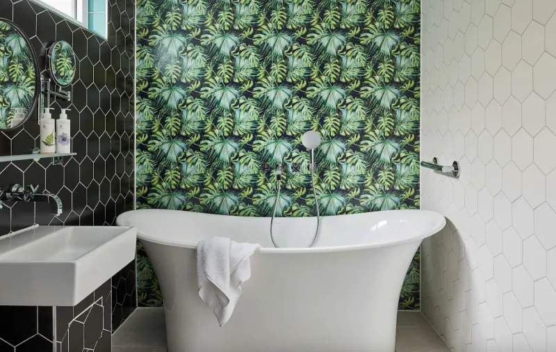 Elegant bathroom with freestanding white bathtub draped in towel, green leaf-patterned wall, black hexagonal tiles, and modern sink.