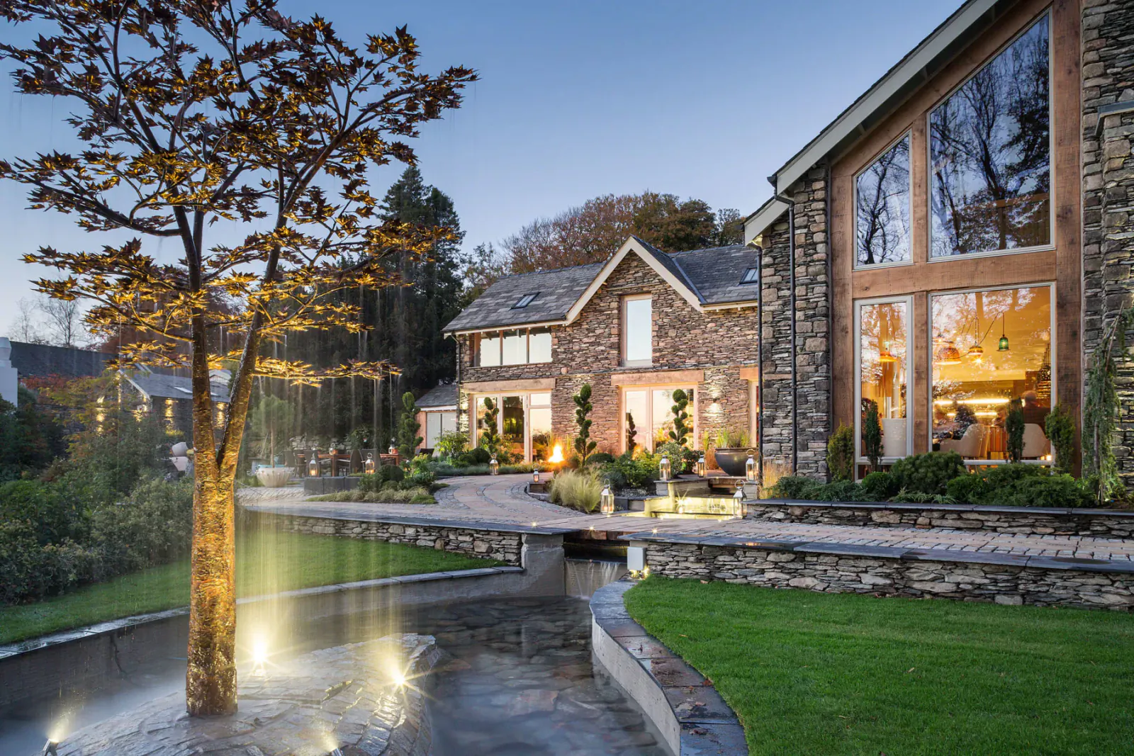 Twilight view of Gilpin Hotel and Lake House in Windermere, Cumbria, with stone buildings, gardens, lit tree, and stream.