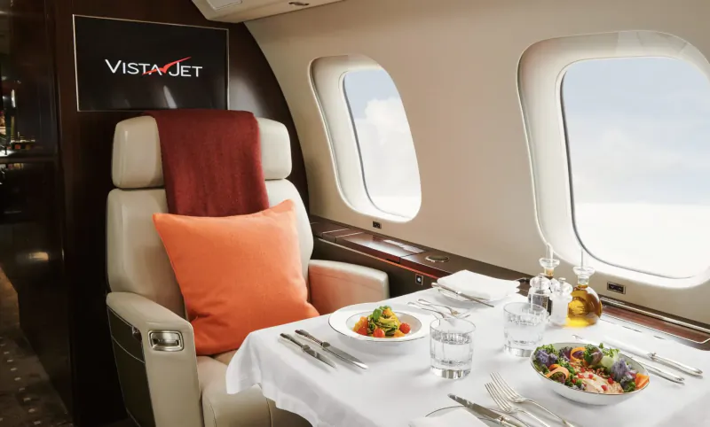VistaJet private jet cabin with luxury beige seat, orange pillow, and healthy salad meal on table by windows.