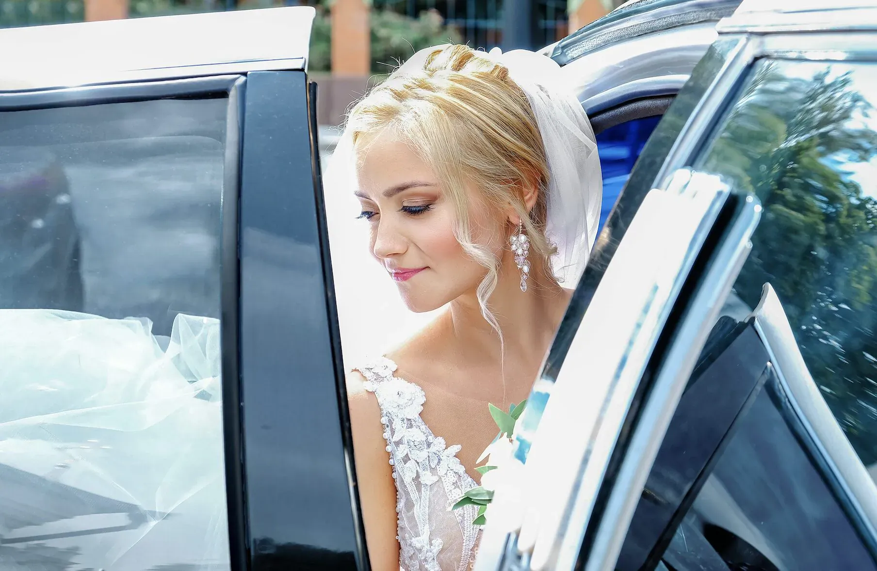 Blonde bride in lace gown and veil steps out of open limousine door on wedding day