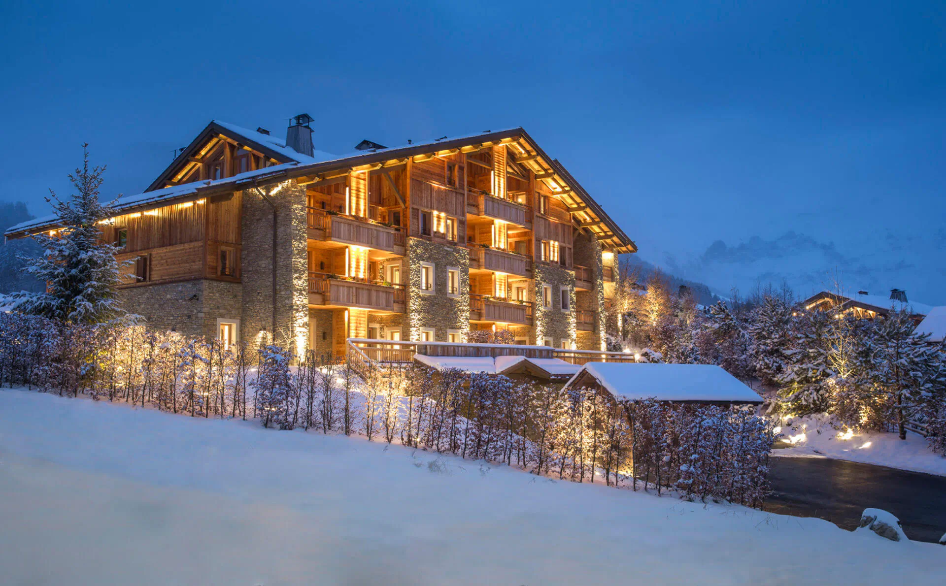 Lit wooden chalet hotel in snowy Megève mountains at dusk, surrounded by snow-covered trees and lights.