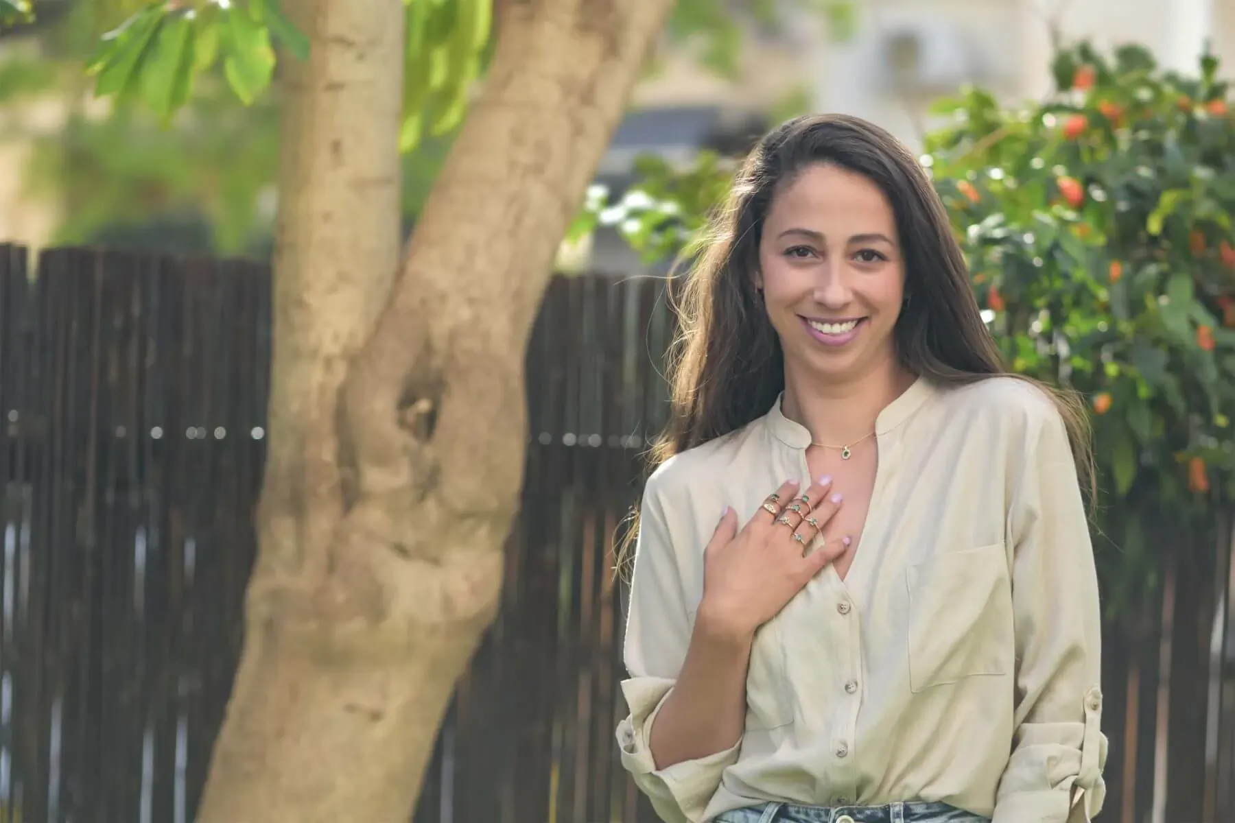 Almog of Mogga Jewels smiling with hand on chest, wearing beige blouse and rings, outdoors by tree and fence