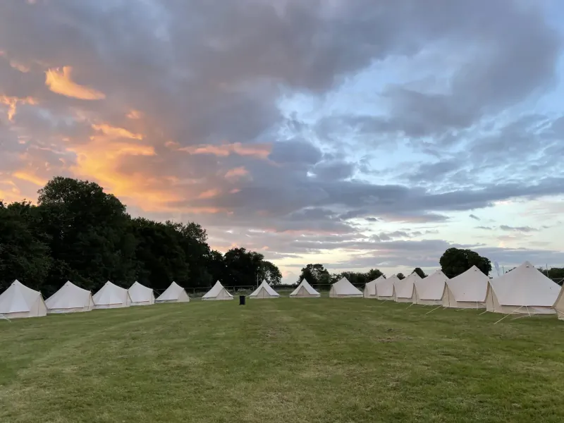 Row of white bell tents on green field at sunset, surrounded by trees under cloudy sky