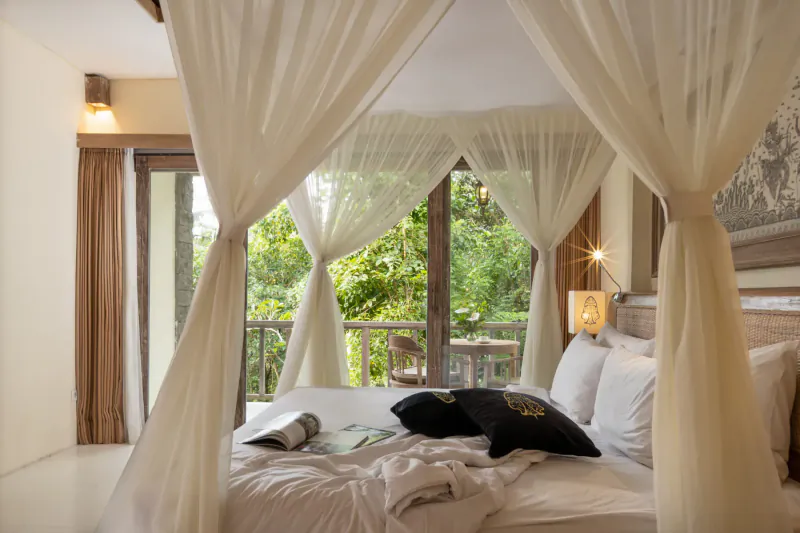 Luxurious canopy bed with white sheer curtains in Kayon Resort Bali suite, lush jungle view through doors.