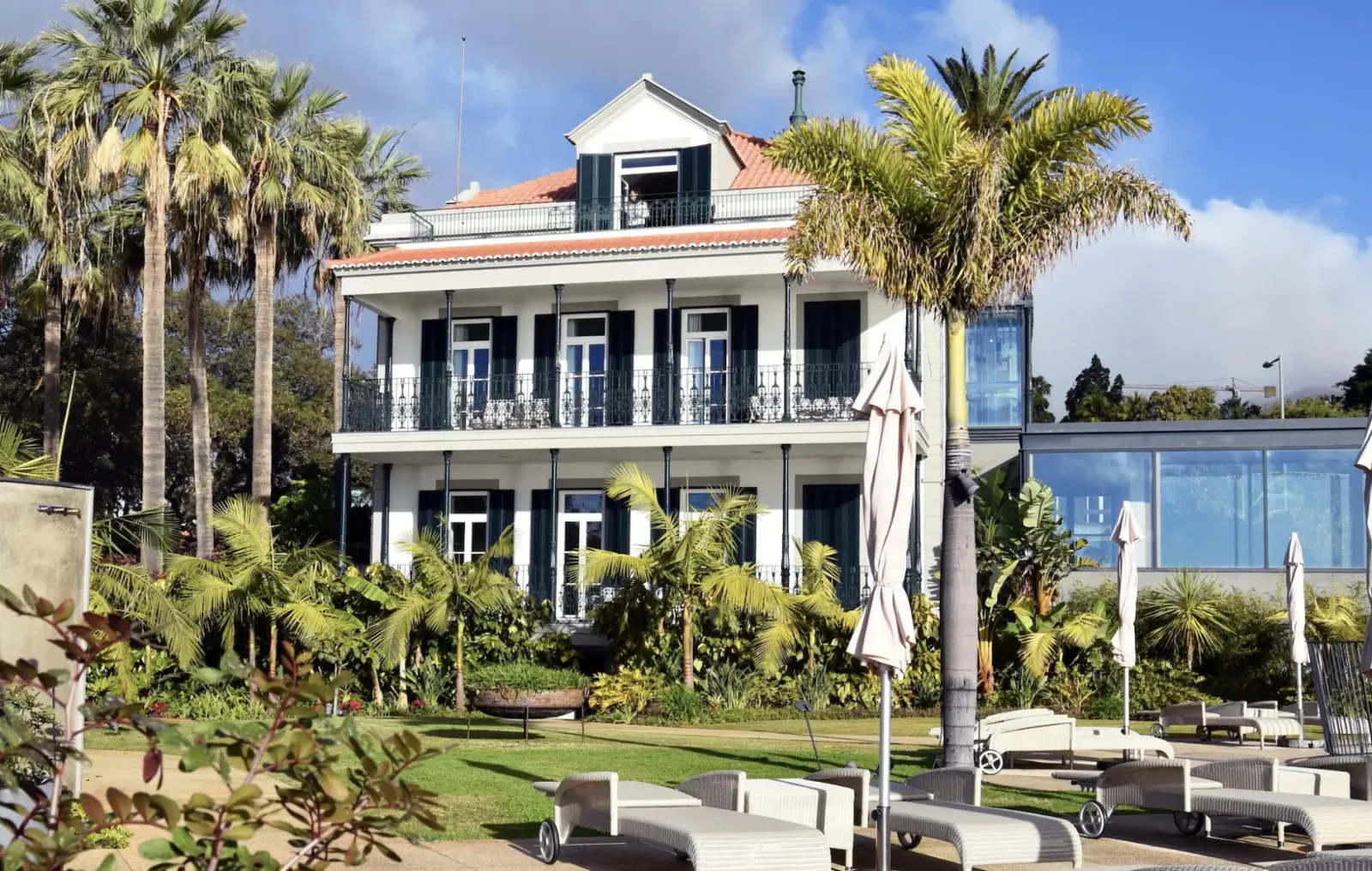 White colonial-style Les Suites Hotel at The Cliff Bay in Funchal, Madeira, with red roof, palms, pool, and loungers.