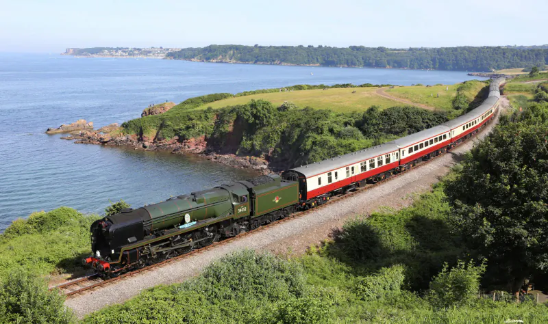 Green steam locomotive pulling luxury coaches along coastal tracks with sea cliffs and green hills.