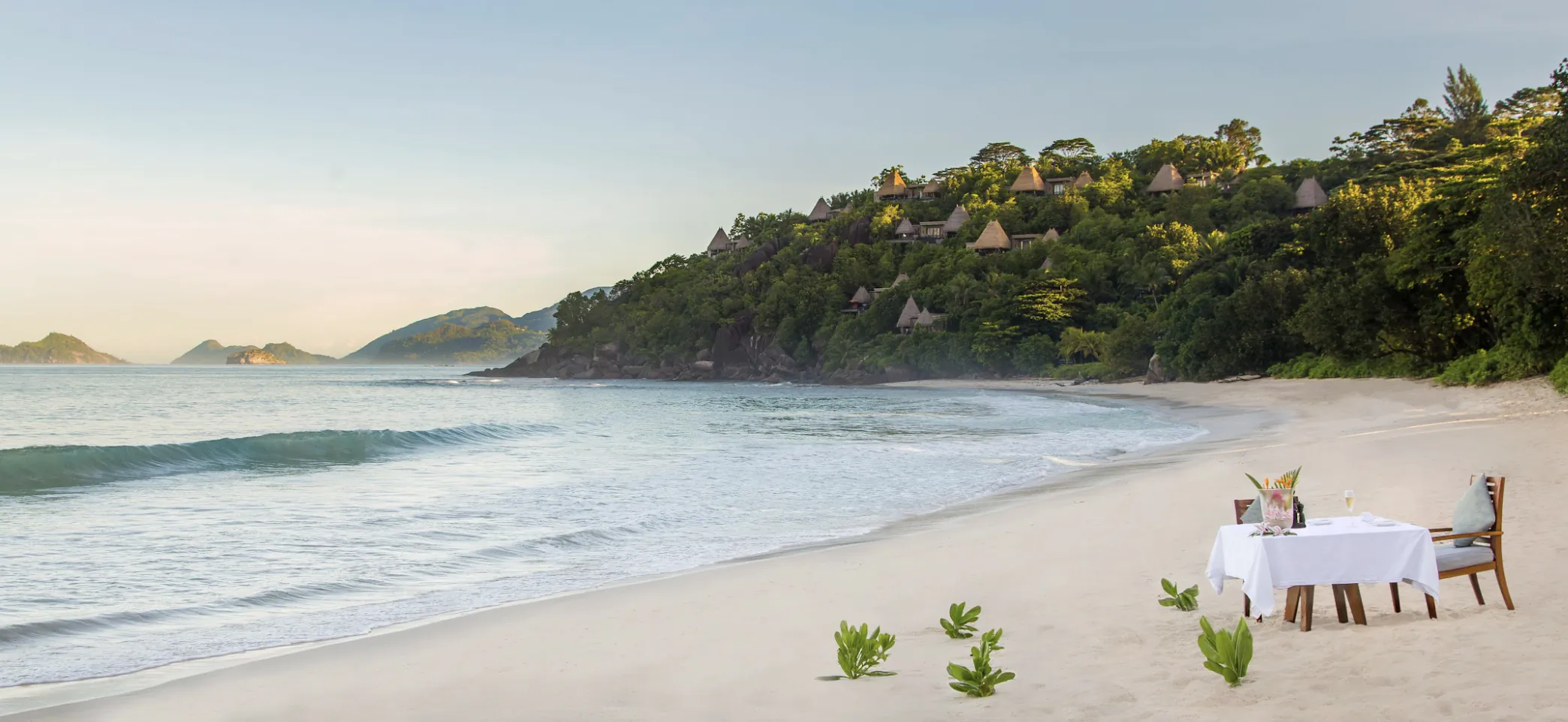 Romantic candlelit dinner table on sandy beach at Anantara Maia Seychelles Villas, with ocean waves, lush green hillside villas, and distant islands at sunset.