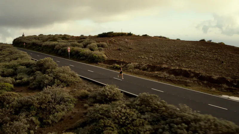 Female triathlete Nicola runs on a winding rural road amid volcanic hills under cloudy skies.