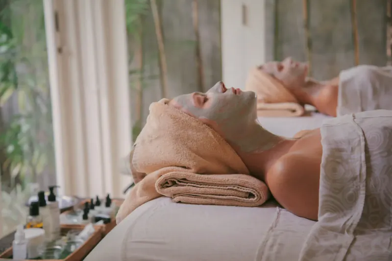 Two women reclining on spa beds with facial masks and towels, surrounded by bamboo and greenery in luxury Bali spa.