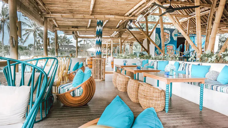 Azul Beach Club Bali: open-air tropical lounge with turquoise cushions, wooden tables, rattan chairs on deck overlooking palms.